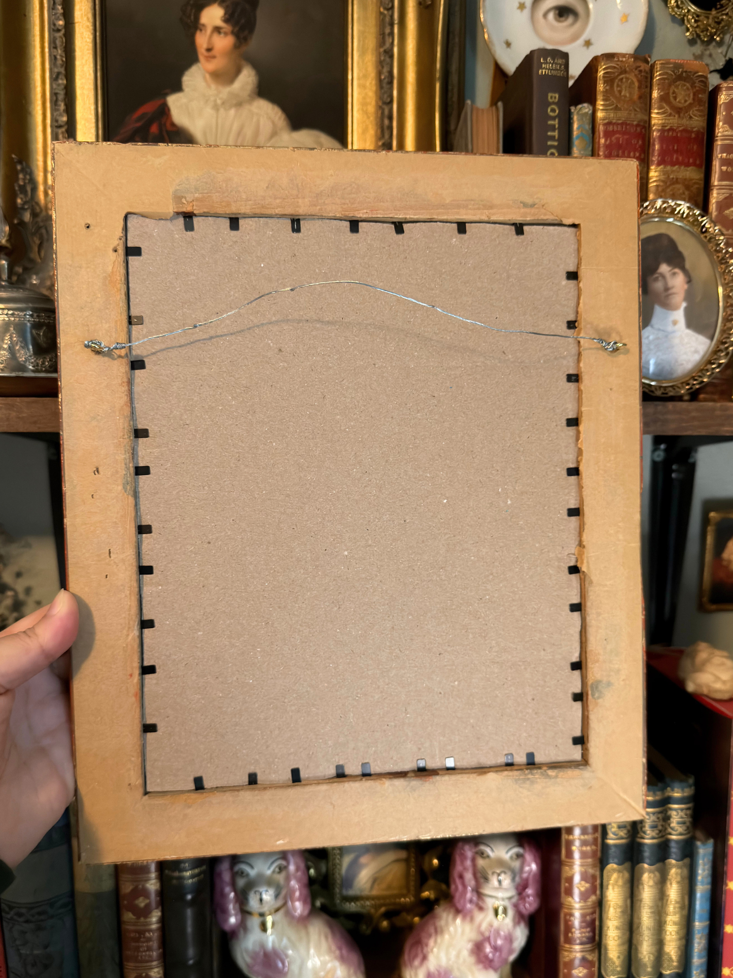 Back view of a framed artwork with a person holding it, surrounded by books and decorative items.