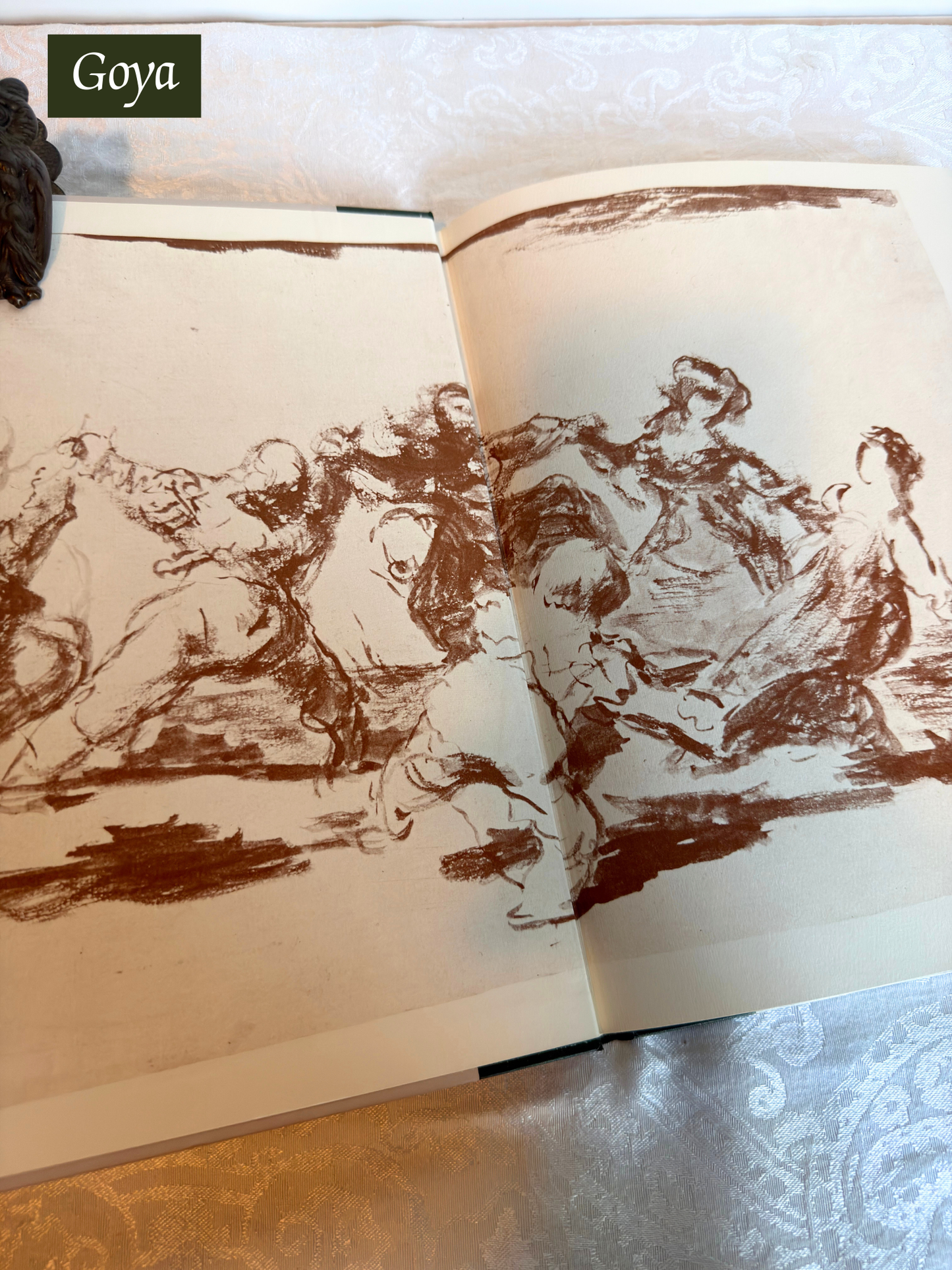 Open book displaying black and white line drawings of figures, with 'Goya' branding at the top.