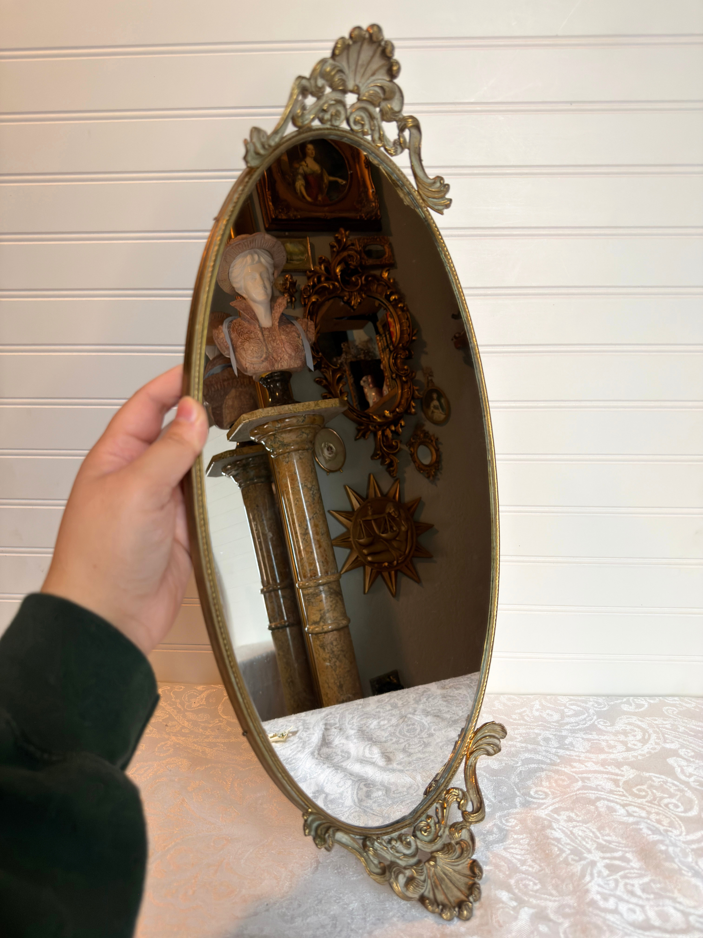 Decorative oval mirror with intricate gold frame held by a hand against a neutral background