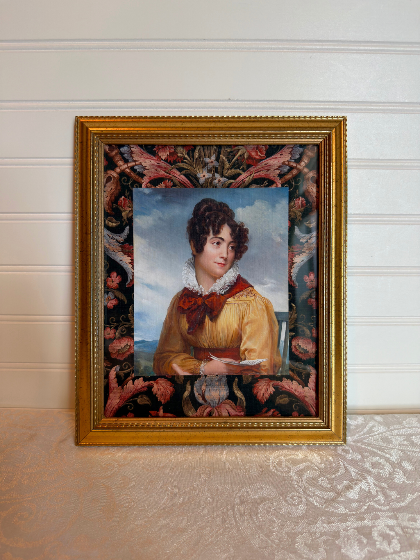 Gold framed portrait of a woman wearing a yellow and red dress holding a book with a decorative floral border. 