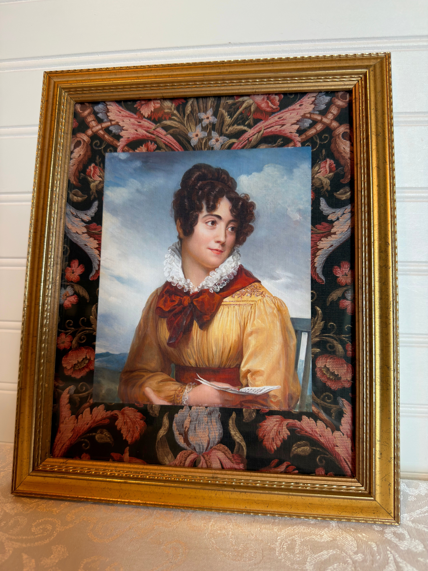 Gold framed portrait of a woman wearing a yellow and red dress holding a book with a decorative floral border