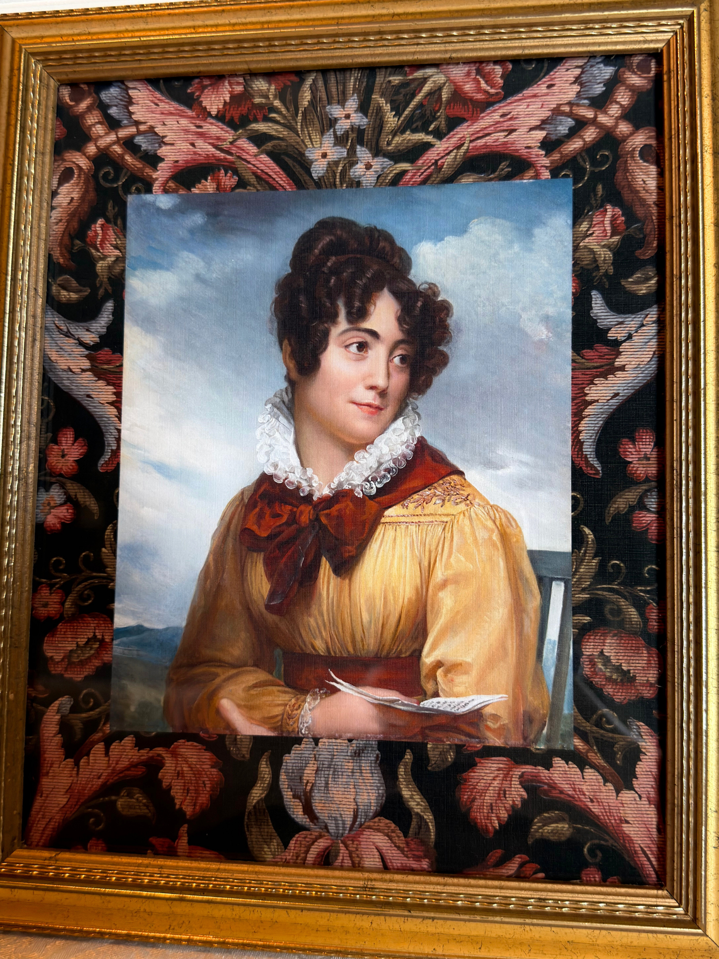Gold framed portrait of a woman wearing a yellow and red dress holding a book with a decorative floral border