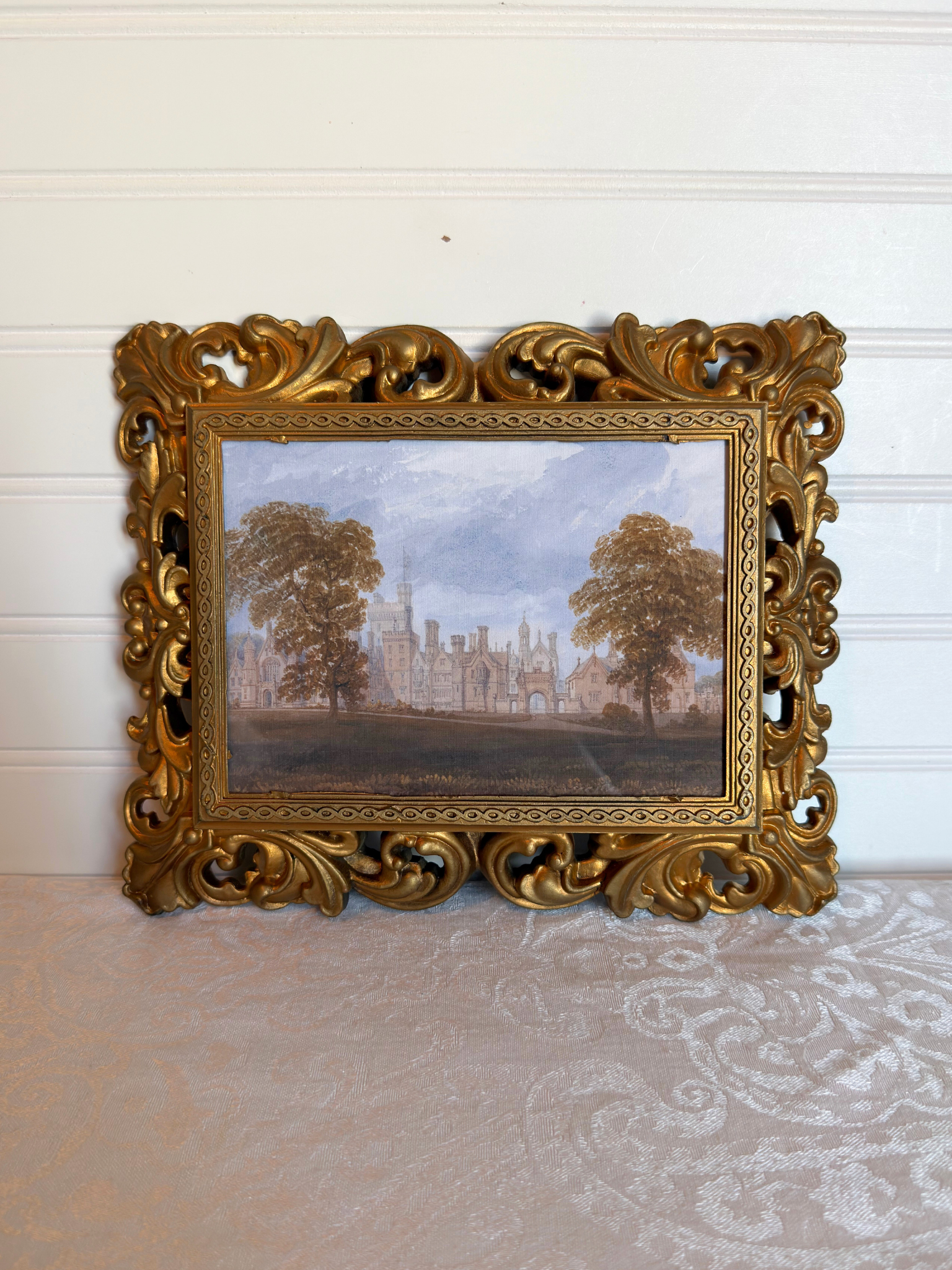Decorative gold frame with a print of a landscape with trees and a manor house against a white background