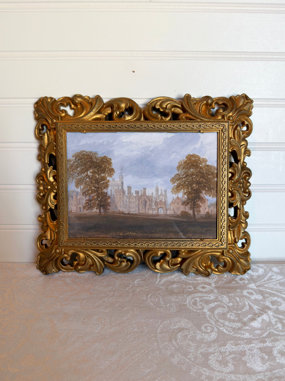 Decorative gold frame with a print of a landscape with trees and a manor house against a white background