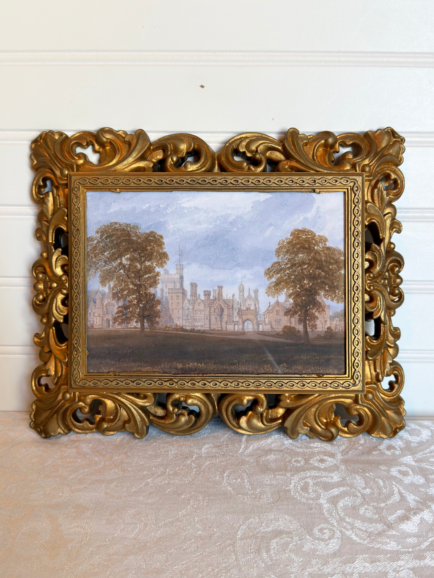 Decorative gold frame with a print of a landscape with trees and a manor house against a white background