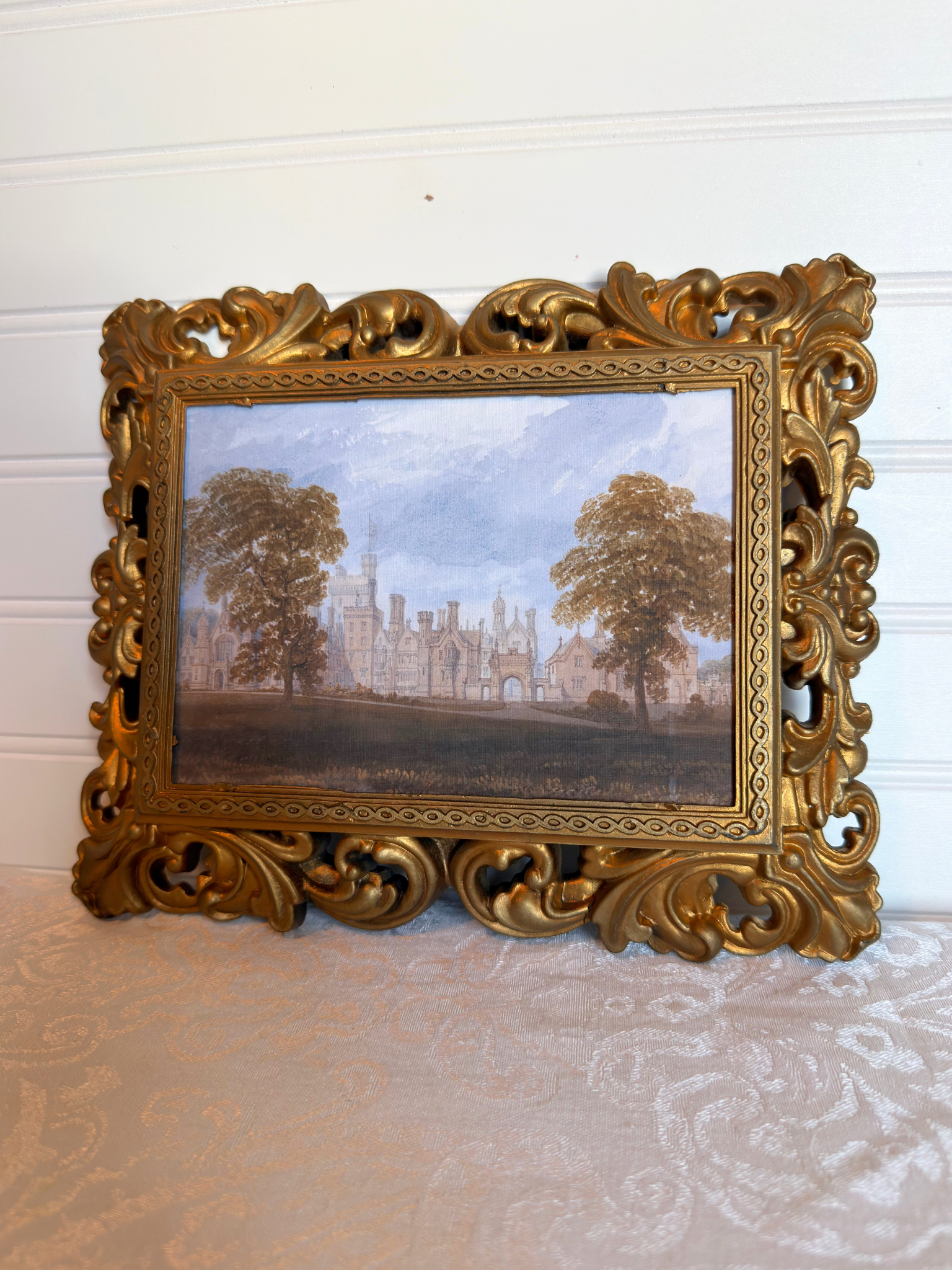 Decorative gold frame with a print of a landscape with trees and a manor house against a white background