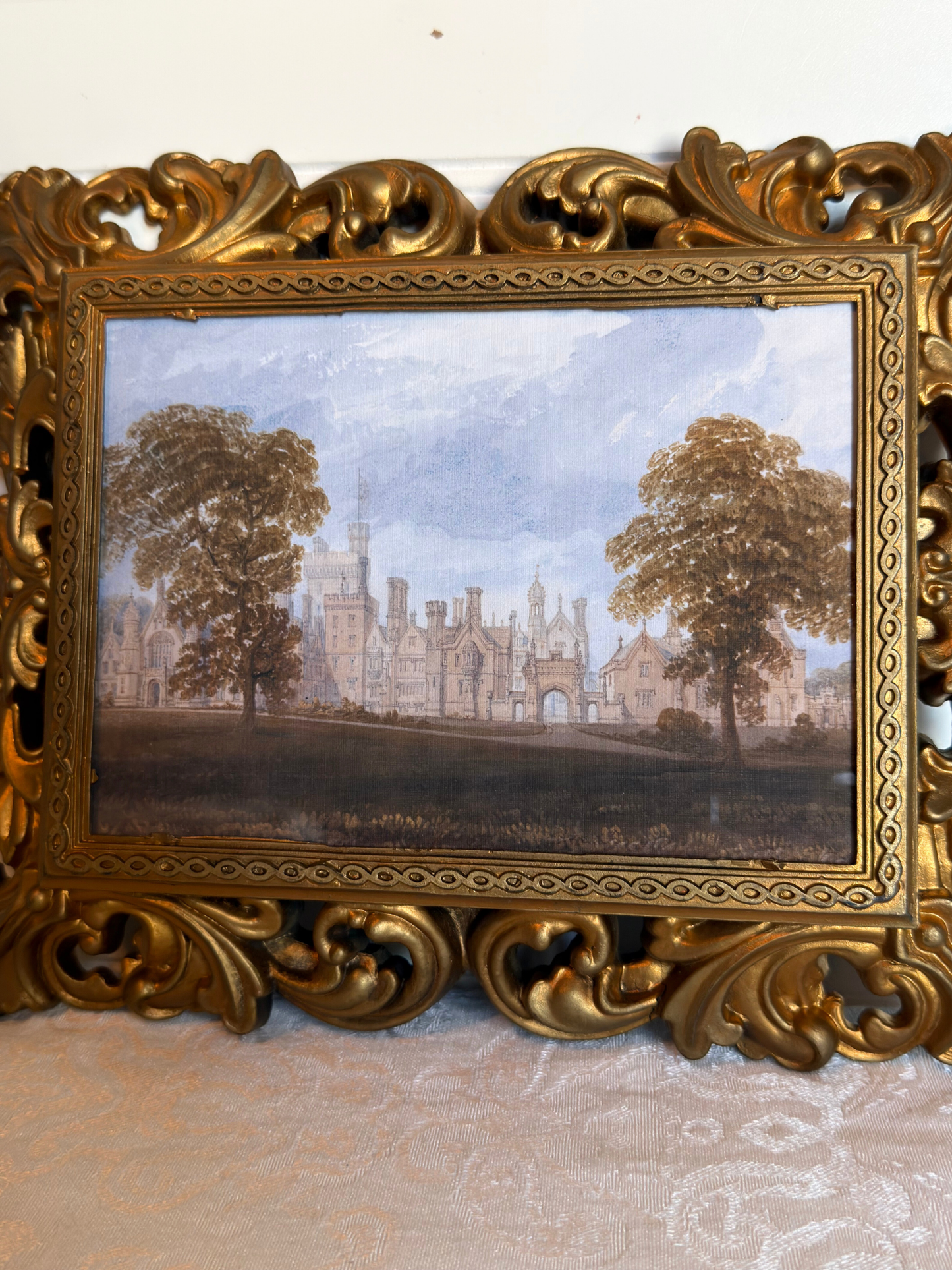 Decorative gold frame with a print of a landscape with trees and a manor house against a white background