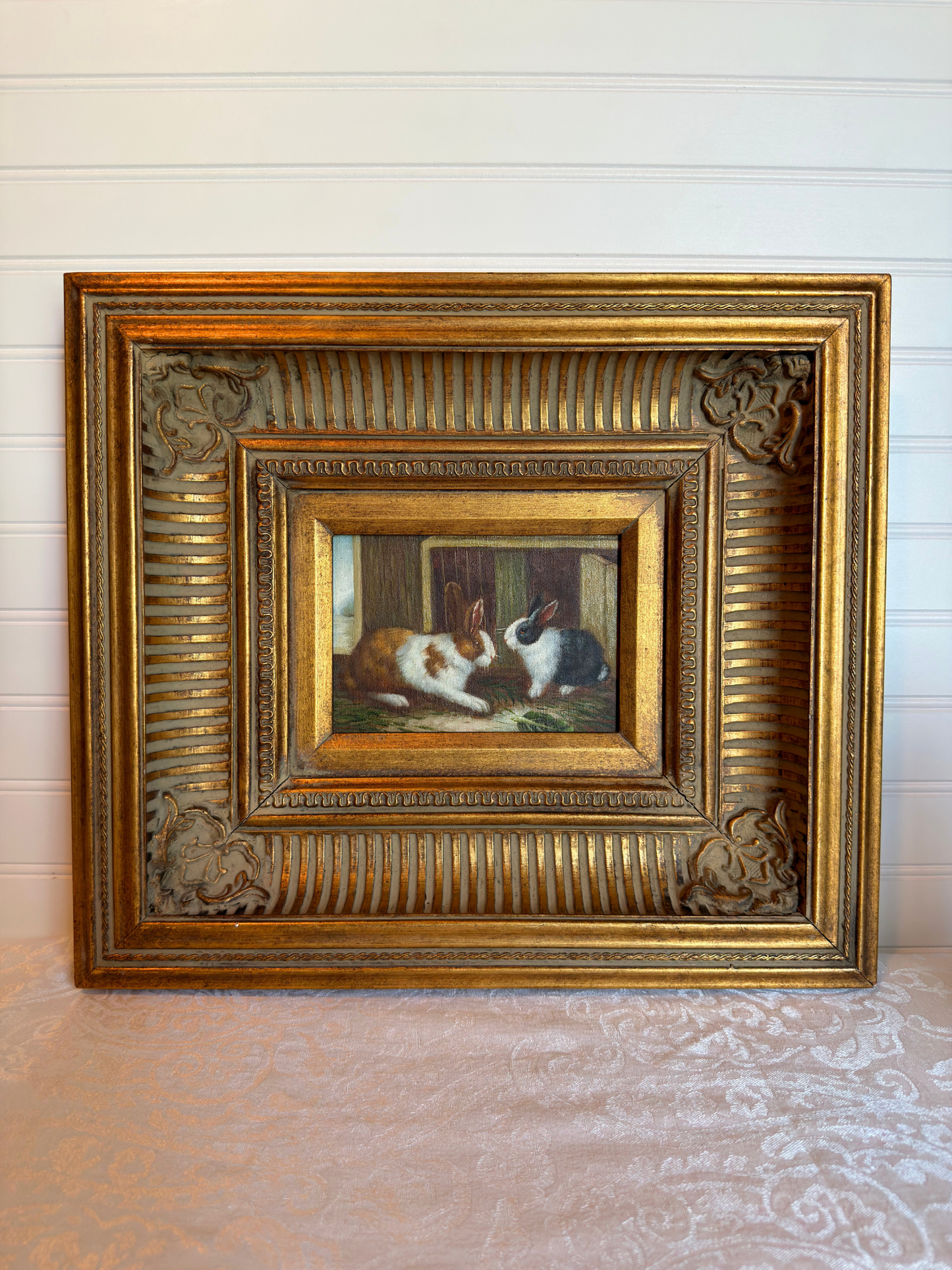 Framed painting of two bunnies in a decorative gold frame on a white background