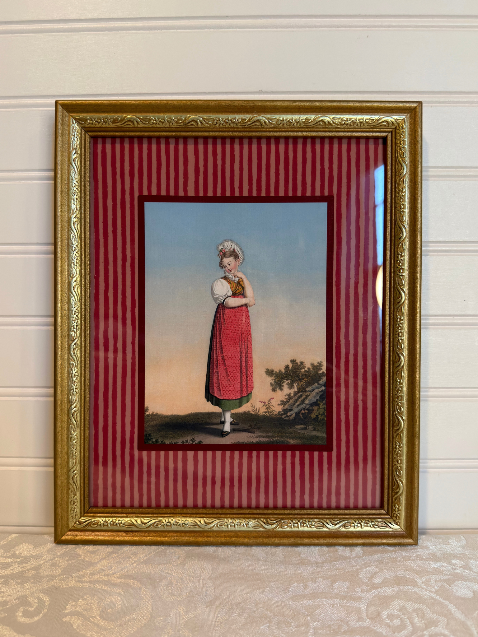 Framed artwork of a woman in a red dress, with a gold frame and red striped inner lining.