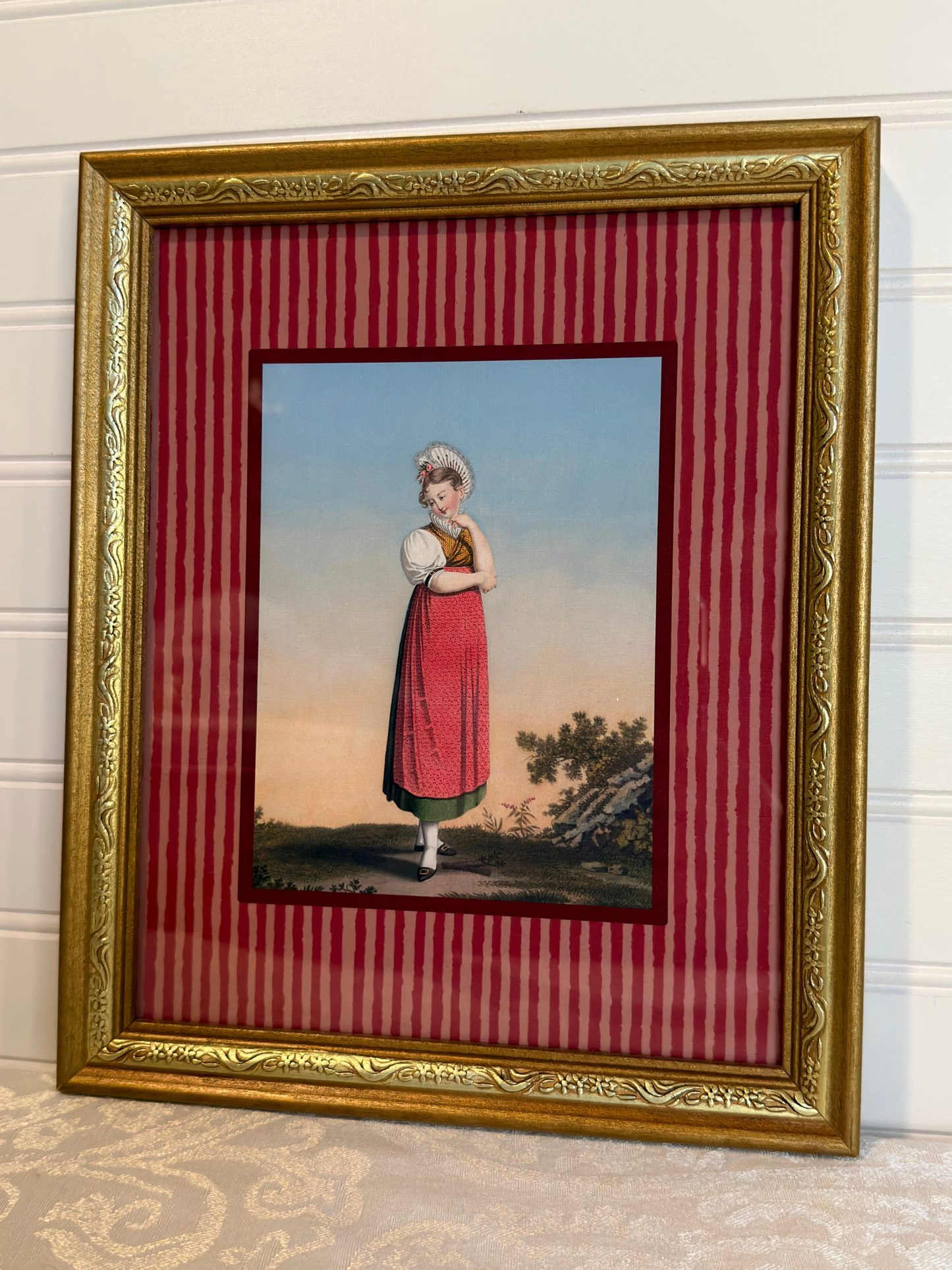 Framed artwork of a woman in a red dress, with a gold frame and red striped inner lining.