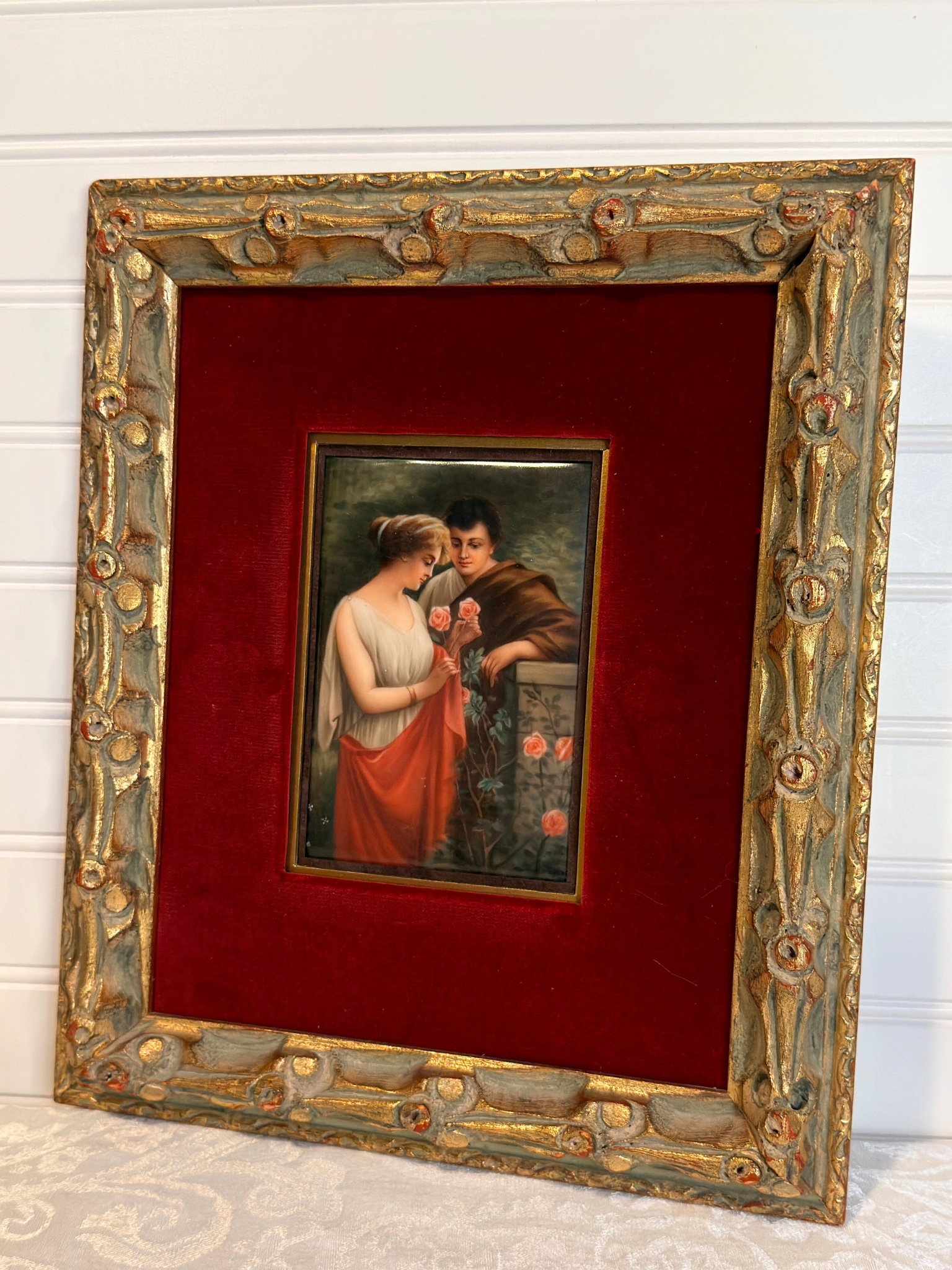 Small painting of couple with roses  within a decorative gold frame on a red background