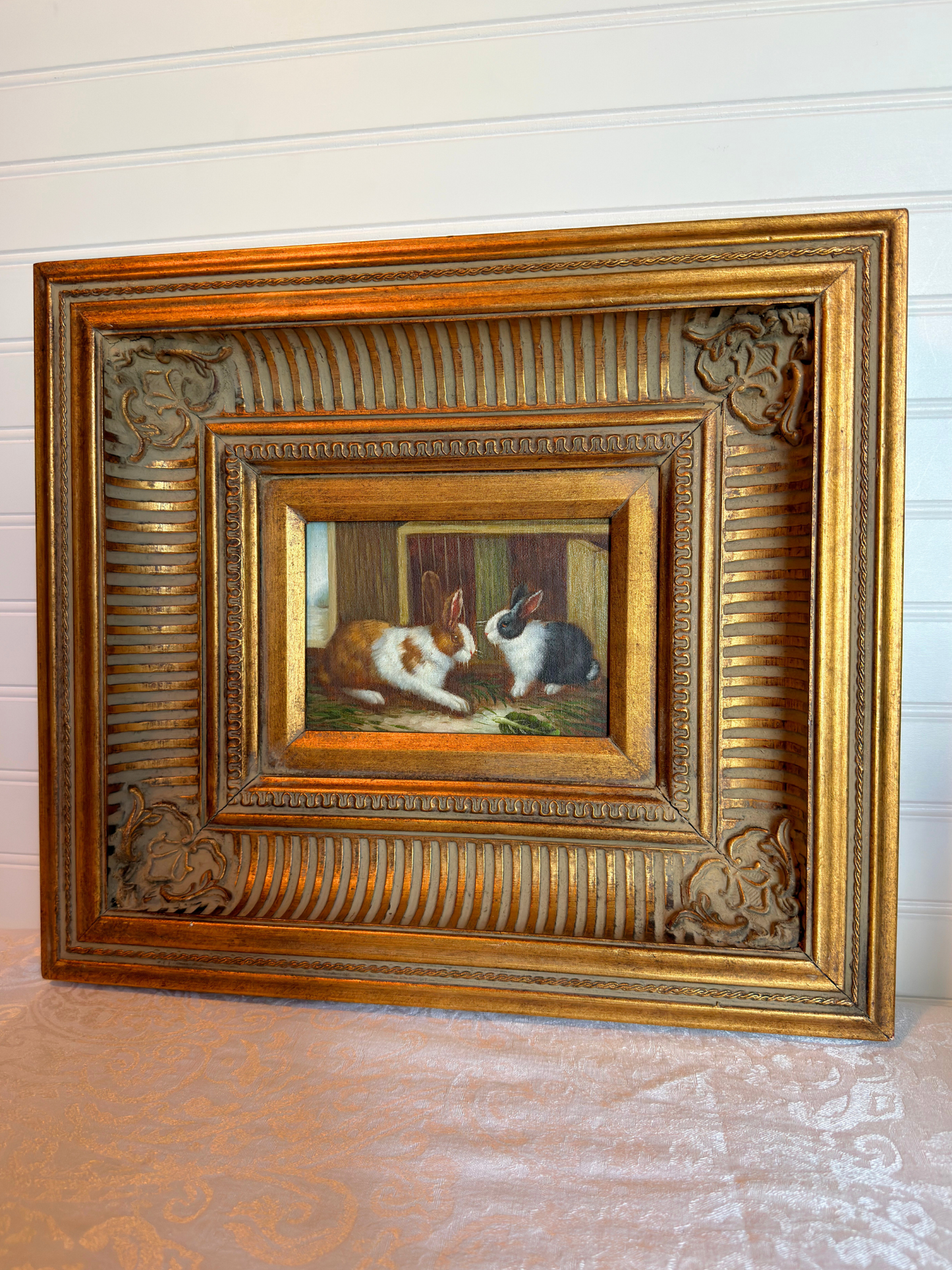 Decorative painting of two rabbits in a ornate gold frame on a textured surface.